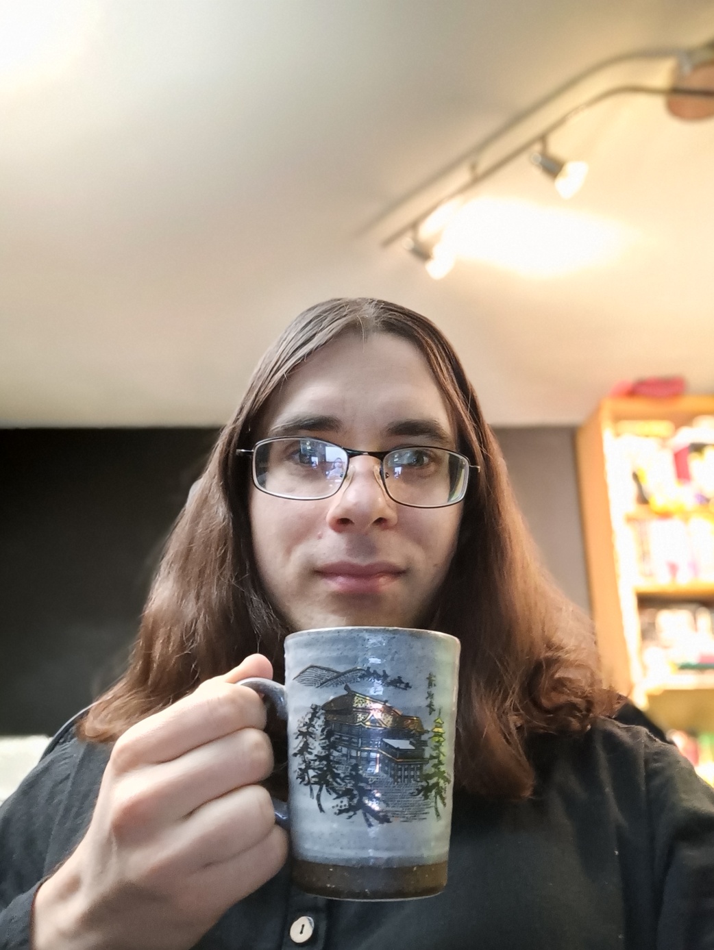 Enby with a cup of tea. On the cup is a picture of a Japanese-style house surrounded by trees. The enby has shoulder-length brown hair and is wearing glasses, which are slightly slanted.